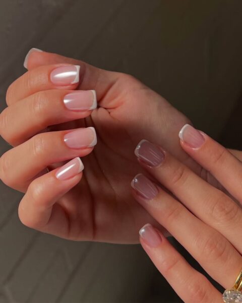 Zdjęcie dłoni z kwadratowymi paznokciami pomalowanymi w stylu french manicure z perłowym wykończeniem. Dłonie pozują tak, by prezentować manicure.