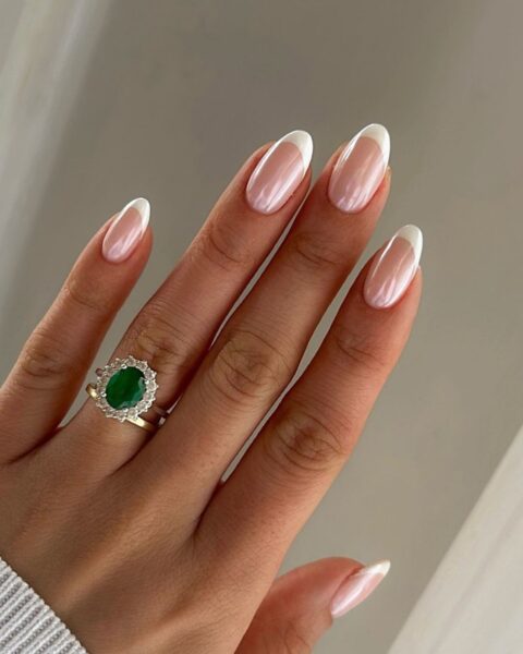 Zdjęcie dłoni z owalnymi paznokciami, które zdobi perłowy french manicure, czyli z perłowym pyłkiem. Na palcu serdecznym znajduje się srebrny pierścionek z zielonym kamieniem.