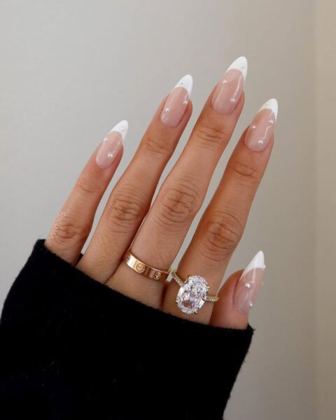 Zdjęcie dłoni z długimi, migdałowymi paznokciami w stylu french manicure z perełkami 3D. Na palcach znajdują się złota obrączka oraz masywny pierścionek z brylantem.