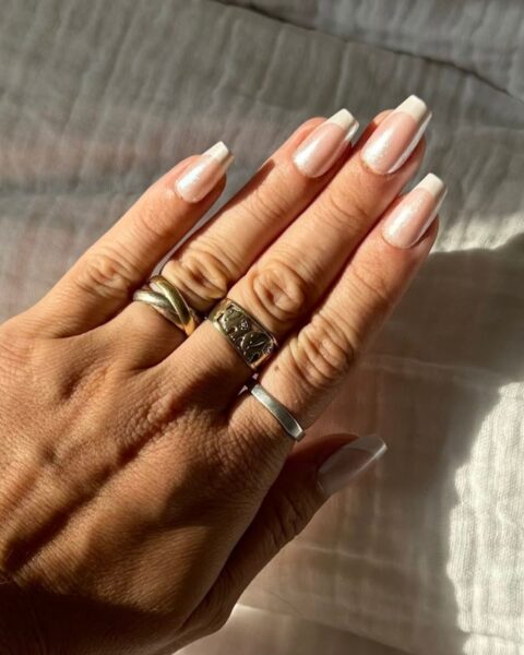 Zdjęcie dłoni z długimi, kwadratowymi paznokciami z french manicure i perłowym połyskiem. Na palcach widać kilka złotych pierścionków. Dłoń pozuje na tle białej pościeli muślinowej.