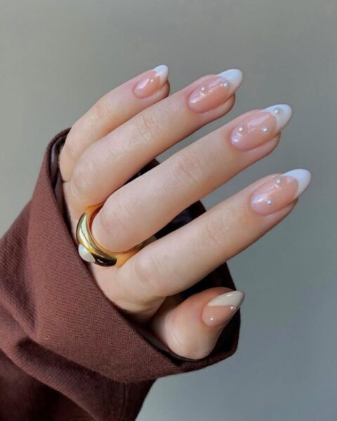Zdjęcie dłoni ze zbliżeniem na długie migdałowe paznokcie w stylu french manicure, ozdobione perełkami 3D. Na palcu środkowym znajduje się masywny, złoty pierścionek z perłą.
