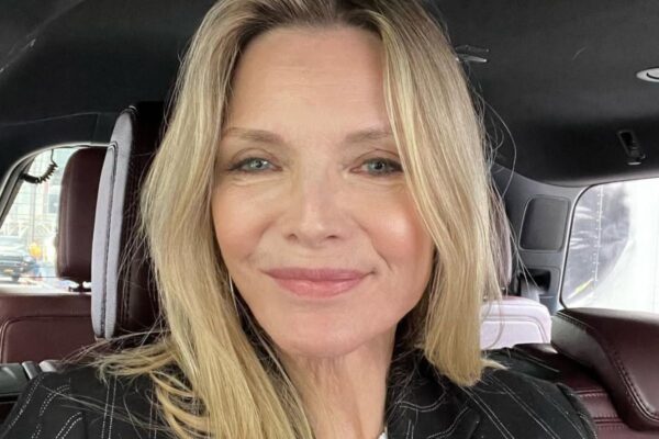 Zdjęcie Michelle Pfeiffer, aktorki, która jest dojrzałą kobietą. Na twarzy ma róż do policzków i bronzer dla 60-latki. Włosy blond, rozpuszczone. Ubrana jest w czarną, prążkowaną marynarkę i siedzi w samochodzie.