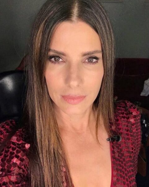 Zdjęcie aktorki Sandry Bullock. Robi sobie selfie na wprost. Ma makijaż z konturem i podkreślonymi oczami. Ubrana jest w cekinową, czerwoną bluzkę z głębokim dekoltem. Ma długie, rozpuszczone, proste włosy. Siedzi na czarnej kanapie.