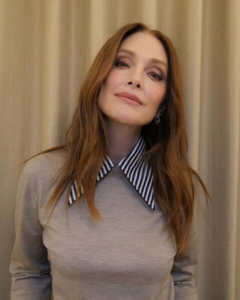 Zdjęcie aktorki Julianne Moore. Ma rude, rozpuszczone na ramiona włosy. Ma podkreślający urodę makijaż z cieniami na powiekach i bronzerem oraz różem na policzkach. Ubrana jest w beżowy sweter, spod którego wystaje kołnierz koszuli w biało-czarne paski. Pozuje na tle beżowej zasłony.