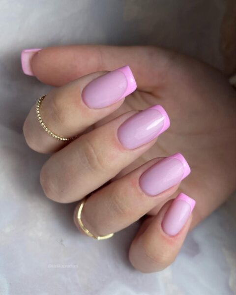 Zdjęcie dłoni ze zbliżeniem na kwadratowe paznokcie z french manicure zrobionego z dwóch kolorów różu. Na palcach znajdują się złote pierścionki. Dłoń pozuje na marmurkowym tle.