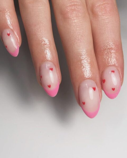Zdjęcie dłoni ze zbliżeniem na długie, migdałowe paznokcie z różową końcówką french manicure oraz ozdobione drobnymi wzorami serduszek. Dłoń pozuje na białym tle.