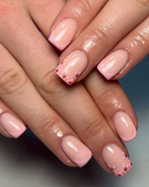 Zdjęcie dłoni ze zbliżeniem na kwadratowe paznokcie w stylu floral french manicure, w którym przy kresce na brzegu paznokcia namalowano kwiatki. Dłonie pozują ułożone jedna na drugą, by prezentować manicure.