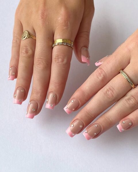 Zdjęcie dłoni ze zbliżeniem na kwadratowe paznokcie. Są pomalowane w stylu french manicure ale z różową końcówką i ozdobione kryształkami. Dłonie pozują na białym blacie.