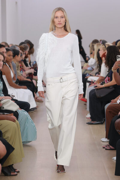 Modelka na pokazie Proenza Schouler SS24 w minimalistycznej letniej stylizacji — białe jeansy o luźnym kroju i lekki, biały sweter z ozdobnym piórem na ramieniu, stylizacja podkreślona prostą fryzurą i naturalnym makijażem.