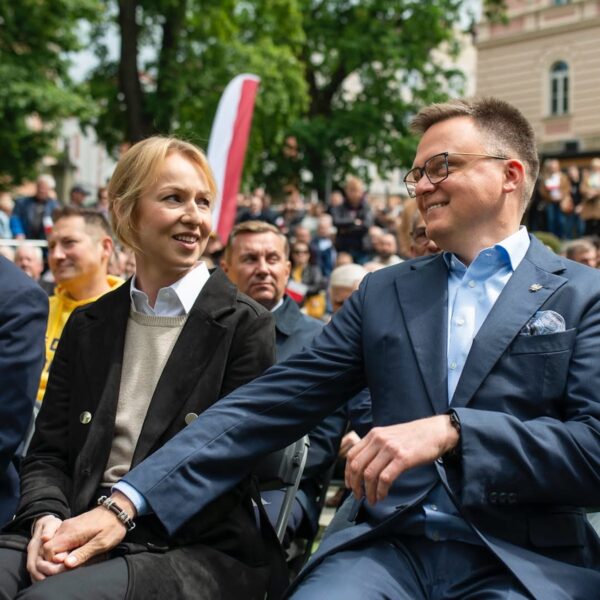 Uśmiechnięta para siedzi obok siebie na wydarzeniu plenerowym, trzymając się za ręce; kobieta w jasnym swetrze i czarnym płaszczu, mężczyzna w granatowym garniturze i okularach, w tle tłum ludzi i polska flaga. Jest to Szymon Hołownia i Urszula Brzezińska-Hołownia.
