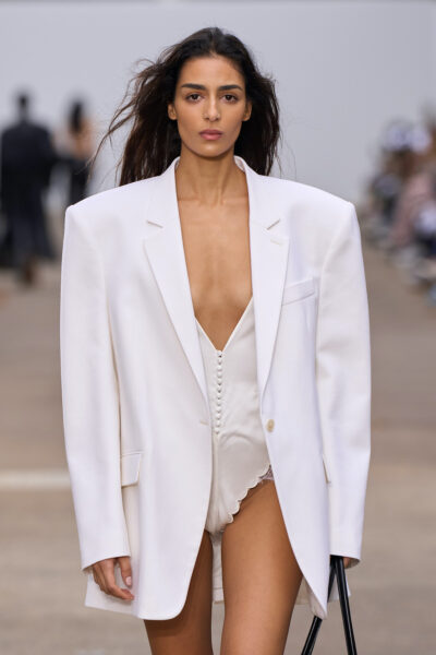 Modelka na wybiegu w oversize’owej białej marynarce i satynowym body z głębokim dekoltem i dekoracyjnym rzędem guziczków. Stylizacja łączy elegancję z bieliźnianą estetyką, podkreślając kontrast form i tkanin.