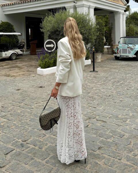 Zdjęcie kobiety na weselu. Ubrana jest w koronkową sukienkę, do tego ma marynarkę oversize w kolorze kremowym. Włosy blond, z półupięciem. W ręce trzyma torebkę Diora. Pozuje naprzeciwko domu weselnego.