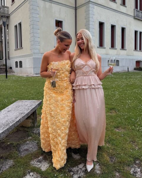 Zdjęcie dwóch kobiet spacerujących w ogrodzie przy pałacu. Mają efektowne stylizacje na wesele. Błędy uniknęły, bo znały zasady. Jedna ma żółtą sukienkę z motywem róż, a druga plisowaną, różową sukienkę i czółenka w szpic. Idą z kieliszkami w dłoni.