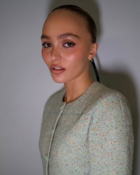 Zdjęcie Lily Rose Depp. Ma włosy gładko związane w kok. Ubrana jest w zieloną, tweedową marynarkę. Makijaż z obrysowanymi na czarno oczami oraz usta obrysowane konturówką. Pozuje na tle ściany.
