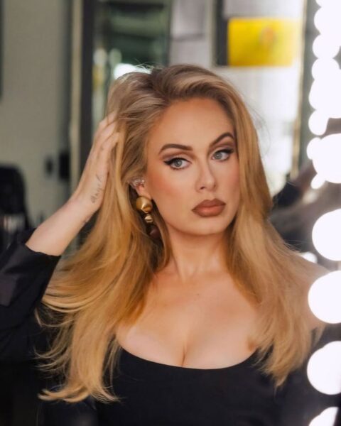 Zdjęcie Adele, która pozuje przed toaletką w lustrem. Poprawia sobie ręką swoje długie, blond włosy. Ma mocny, sceniczny makijaż z podkreślonymi oczami oraz ustami podkreślonymi konturówką. Ubrana jest w czarną sukienkę z głębokim dekoltem.