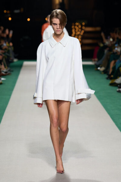 Modelka w oversize’owym, białym płaszczu z szerokim kołnierzem i mankietami, noszonym jako sukienka. Stylizacja minimalistyczna, bez widocznych dodatków, prezentowana na zielono-białym wybiegu.