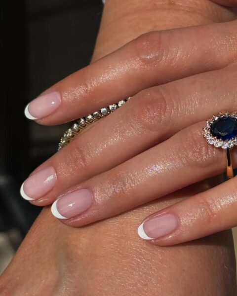 Zdjęcie dłoni ze zbliżeniem na paznokcie z vintage french manicure. Dłoń opiera się o drugą rękę, by prezentować manicure. Widać bransoletkę wysadzaną cyrkoniami oraz pierścionek z niebieskim kamieniem.