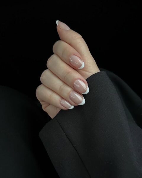 Zdjęcie dłoni ze zbliżeniem na paznokcie ze stylizacją french manicure. Dłoń pozuje na czarnym tle. Widać fragment rękawa czarnej marynarki.