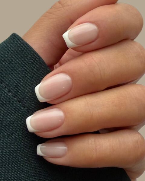 Zdjęcie dłoni z długimi, kwadratowymi paznokciami pomalowanymi w stylu french manicure. Palce dłoni zwijają się na czarnym rękawie bluzki.