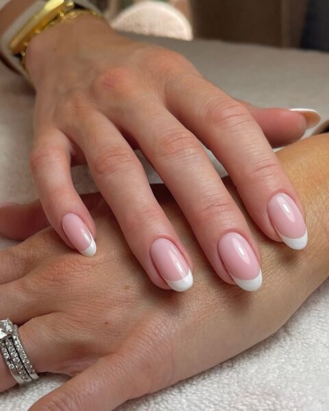 Zdjęcie dłoni ze zbliżeniem na paznokcie. Zdobi je french manicure. Dłoń układa się na drugiej ręce i prezentuje manicure. Widać w tle złoty zegarek, a także srebrny pierścionek z brylantem na palcu na pierwszym planie. Dłonie pozują na ręczniku, tuż po stylizacji.