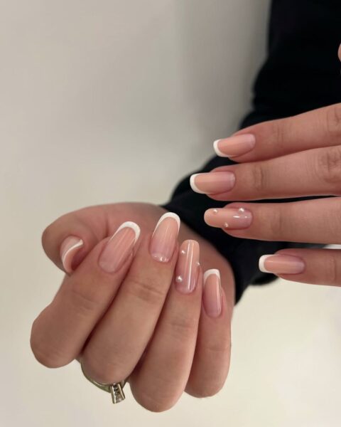 Zdjęcie dłoni ze zbliżeniem na długie, owalne paznokcie. Zdobi je french manicure oraz kilka perełek. Dłonie pozuje na tle białej ściany tak, by prezentować manicure. Na jednym z palców znajduje się srebrny pierścionek z kryształkiem.