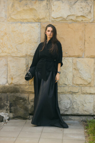 Kobieta w czarnej kimono-podobnej bluzce z paskiem i długiej, błyszczącej spódnicy, stoi przy kamiennej ścianie, trzymając dużą czarną torebkę typu kopertówka.