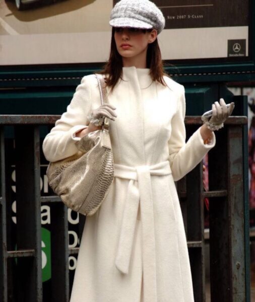 Kobieta w eleganckim, białym płaszczu z paskiem w talii, trzymająca srebrną torbę na ramieniu i telefon w ręku, stoi na tle wejścia do stacji metra. Ma na sobie szare rękawiczki i modną czapkę z daszkiem. Jest to kadr z filmu "Diabeł ubiera się u Prady".