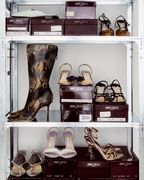 Metalowy regał wypełniony eleganckimi butami Jimmy Choo – szpilki, sandały i kozaki w różnych stylach i kolorach, wraz z markowymi pudełkami ułożonymi na półkach. Jest to nowa kolekcja marki inspirowana archiwalnymi projektami.