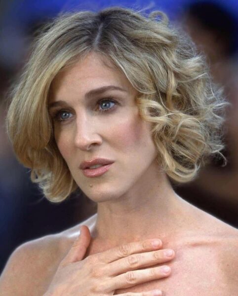 Zbliżenie na kobietę z blond, kręconymi włosami, patrzącą z powagą, z ręką przyłożoną do klatki piersiowej. Jest to Sarah Jessica Parker.