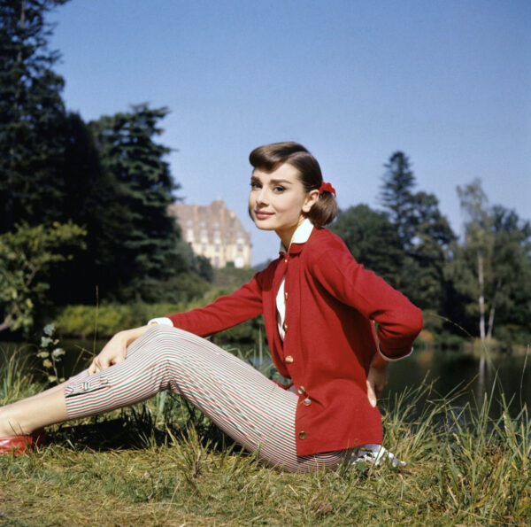 Kobieta Audrey Hepburn siedząca na trawie nad brzegiem jeziora w słoneczny dzień. Ma na sobie czerwony kardigan z ozdobnymi guzikami, białą bluzkę i obcisłe, prążkowane legginsy capri w biało-czerwono-zielone paski. Jej włosy są spięte z kokardką, a w tle widoczne są drzewa i pałac. Stylizacja i kolorystyka nawiązują do mody lat 50. i stylu retro.