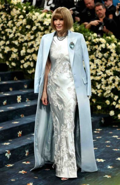 Anne Wintour na MET Gali w 2025 roku. Kobieta z krótkim bobem pozuje na schodach wyłożonych niebieskim dywanem w eleganckiej, srebrzystej sukni z haftem kwiatowym i jasnoniebieskim płaszczem zarzuconym na ramiona; jej stylizację uzupełnia duża broszka i perłowy naszyjnik, w tle widać dekorację z białych kwiatów i fotografów.