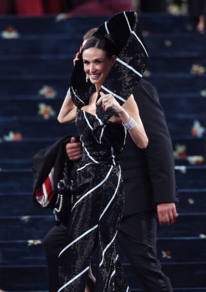 Demi Moore na MET Gali w 2025 roku. Kobieta ubrana w efektowną, czarno-białą, połyskującą suknię z dużym, rzeźbiarskim kołnierzem na czerwonym dywanie; uśmiecha się i trzyma dłonie przy głowie, w tle widać schody oraz ochroniarzy.