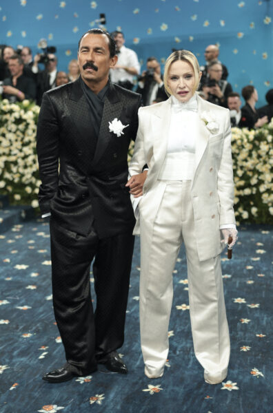 Haider Ackermann oraz Madonna na MET Gali w 2025 roku. Para pozująca na niebieskim dywanie w eleganckich garniturach: mężczyzna w czarnym, satynowym garniturze z białym kwiatem w butonierce oraz kobieta w białym, oversize'owym garniturze z muchą i koronkową rękawiczką, w tle tłum fotografów i kwiatowa dekoracja.