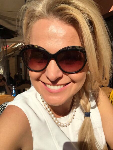 Uśmiechnięta kobieta w dużych okularach przeciwsłonecznych, perłowym naszyjniku i białej bluzce robi selfie w słoneczny dzień, jej blond włosy spięte są w bok w warkocz. Styl Irminy Ochockiej, partnerki Marka Jakubiaka.