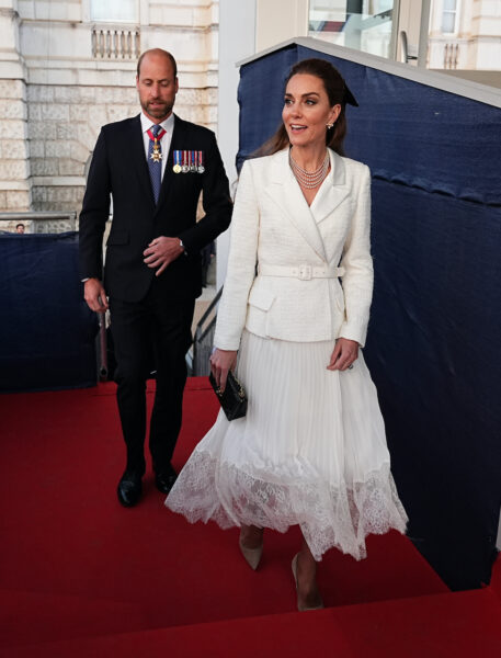 Książę William i księżna Kate wchodzą po czerwonym dywanie na uroczystość. Kate ma na sobie biały żakiet i plisowaną spódnicę z koronką oraz beżowe szpilki, a William jest w ciemnym garniturze z medalami.