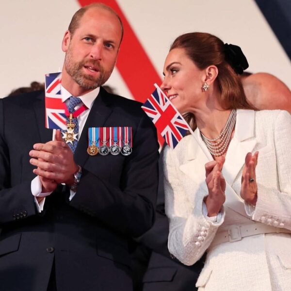 Książę William i księżna Kate Middleton stoją razem, uśmiechnięci i trzymają brytyjskie flagi podczas oficjalnego wydarzenia. William ma na sobie garnitur z odznaczeniami wojskowymi, a Kate ubrana jest w biały kostium i perłowy naszyjnik.