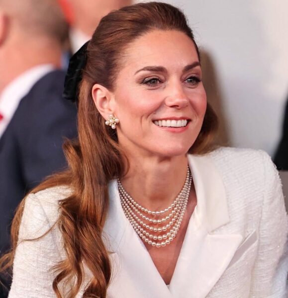 Zbliżenie na uśmiechniętą księżną Kate w białym żakiecie, ozdobioną wielorzędowym perłowym naszyjnikiem i kolczykami z perłami i diamentami. Jej długie włosy są częściowo upięte wstążką. Kate Middleton Dzień Zwycięstwa w Europie i 80. rocznica zakończenia II wojny światowej.