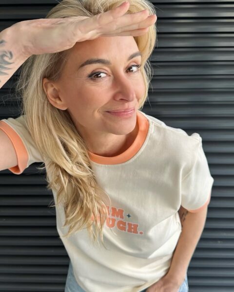Kobieta z długimi blond włosami uśmiecha się i zasłania czoło ręką jakby spoglądała w dal, ubrana w kremowy T-shirt z pomarańczowymi wykończeniami. Jest to dziennikarka i podróżniczka Martyna Wojciechowska dzisiaj.