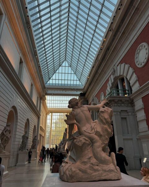 Wnętrze muzeum Metropolitan Museum of Art (Met) w Nowym Jorku z przeszklonym dachem w kształcie trójkąta, oświetlone naturalnym światłem. Na pierwszym planie duża rzeźba przedstawiająca nagą kobietę z wężowym ogonem, siedzącą na skale. W tle widoczne inne klasyczne rzeźby oraz zwiedzający spacerujący po galerii.