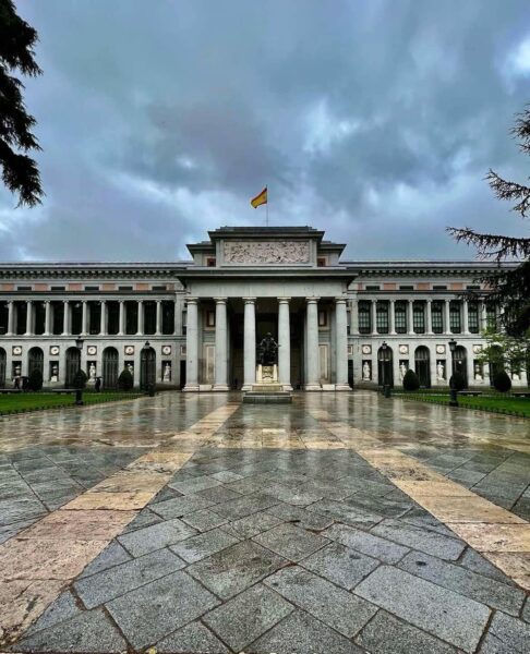 Monumentalna fasada Muzeum Prado w Madrycie, z kolumnowym portykiem, hiszpańską flagą powiewającą na dachu i posągiem przed wejściem. Przed budynkiem mokry, kamienny dziedziniec po deszczu, otoczony trawnikiem i drzewami. Na niebie ciemne, burzowe chmury.