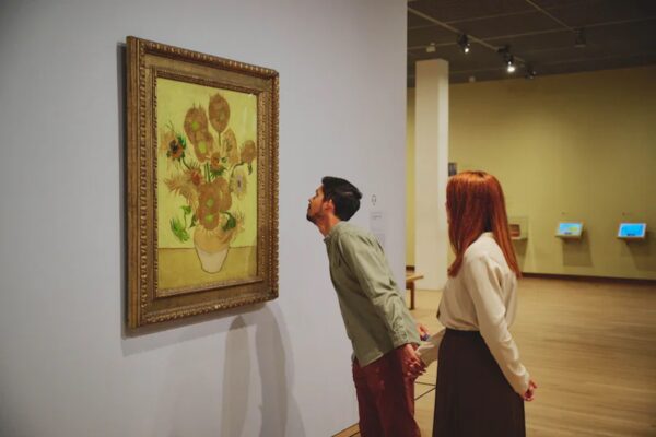 Para zwiedzających ogląda z bliska słynny obraz „Słoneczniki” Vincenta van Gogha w muzeum. Mężczyzna nachyla się, by przyjrzeć się szczegółom, podczas gdy kobieta trzyma go za rękę. Obraz wisi na jasnej ścianie w eleganckiej ramie.