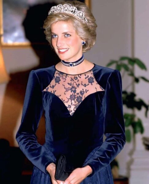 Księżna Diana w eleganckiej granatowej sukni z koronkowym wykończeniem, nosząca diamentową tiarę i choker z szafirem, uśmiechająca się podczas oficjalnego wydarzenia.