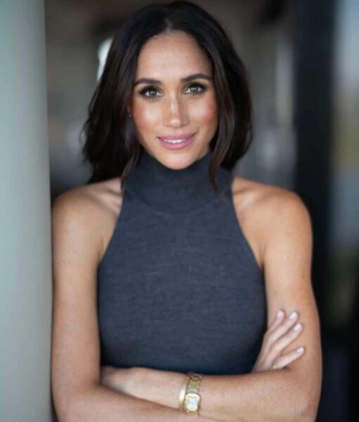 Meghan Markle w bezrękawnikowej, ciemnoszarej bluzce, z rozpuszczonymi włosami i złotym zegarkiem na ręku, uśmiechająca się z założonymi rękami.