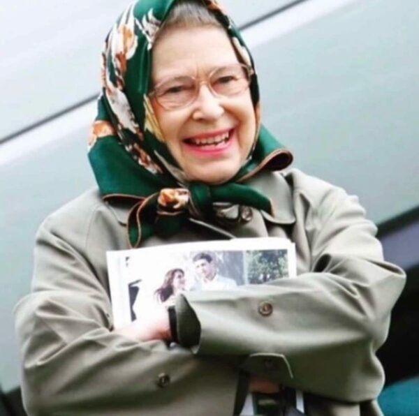 Królowa Elżbieta II uśmiechnięta, ubrana w oliwkowy płaszcz i zieloną chustę w kwiaty, trzymająca złożony magazyn z fotografią młodej pary.