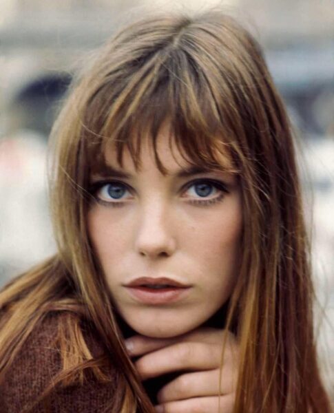 Jane Birkin z długimi, rozpuszczonymi włosami i charakterystyczną grzywką, spoglądająca intensywnie w obiektyw, opierając dłonie pod brodą.