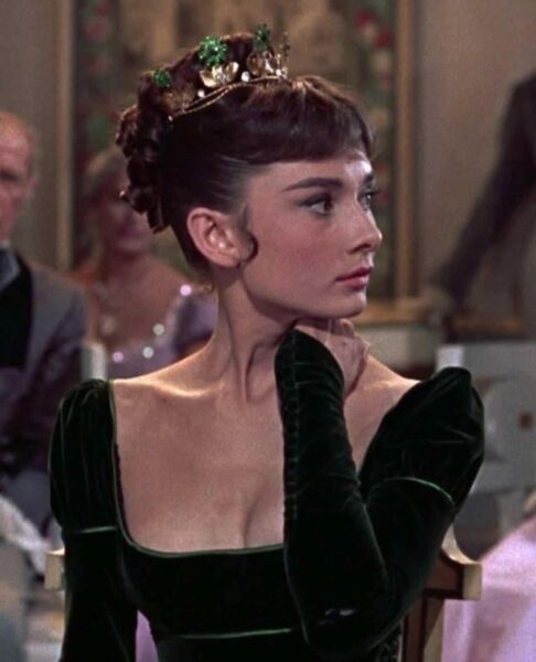 Audrey Hepburn w eleganckiej, aksamitnej sukni z głębokim dekoltem i koroną na głowie, siedząca bokiem w wysmakowanym wnętrzu.