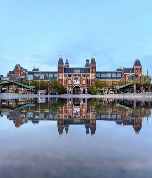 Fasada Rijksmuseum w Amsterdamie, neogotycki budynek z czerwonej cegły z wieżyczkami i łukowym wejściem, odbijający się symetrycznie w spokojnej tafli wody na pierwszym planie. Po bokach widoczne pawilony i drzewa, całość w świetle dnia pod błękitnym niebem.