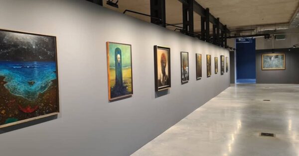 Wnętrze galerii sztuki z szarymi ścianami i obrazami w stylu surrealistycznym zawieszonymi wzdłuż jednej ze ścian; widoczna minimalistyczna, nowoczesna przestrzeń wystawiennicza. Są to obrazy Beksińskiego na wystawie w Warszawie.