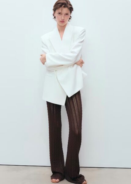 Modelka ubrana w oversize’ową, białą marynarkę zestawioną z ciemnobrązowymi, ażurowymi spodniami z pionowym wzorem. Stylizacja minimalistyczna, utrzymana w neutralnych barwach, idealna na lato w wersji smart casual.