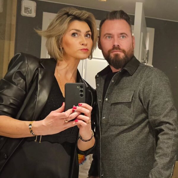 Kobieta i mężczyzna pozują do wspólnego selfie w lustrze; kobieta z blond fryzurą w skórzanej kurtce trzyma telefon, mężczyzna w szarej koszuli patrzy w obiektyw, oboje w eleganckich stylizacjach. Styl Marty Sosnowskiej, żony Krzysztofa Stanowskiego.
